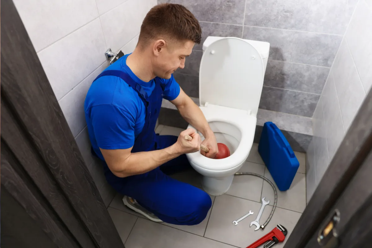 Plumber using rubber plunger to unclog toilet with wrenches and toolbox on bathroom floor